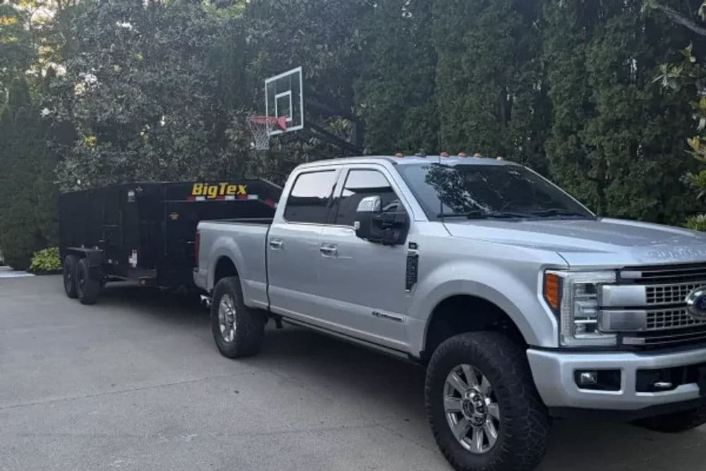 Silver Ford truck towing Big Tex trailer with basketball hoop in driveway surrounded by trees.